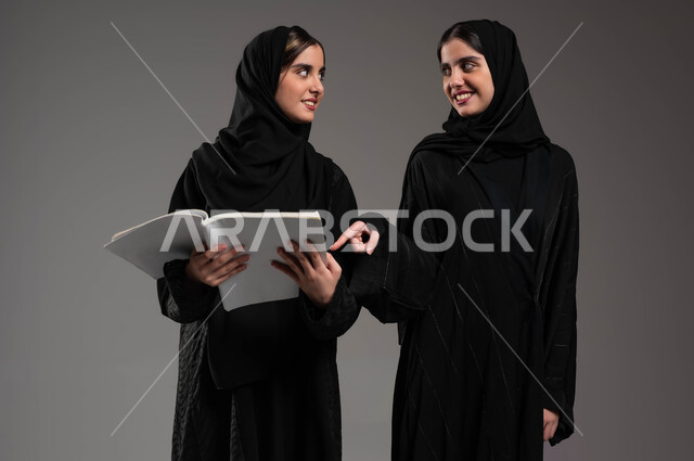 مراجعة الدروس اليومية، الاستمتاع بممارسة هواية المطالعة، قضاء الوقت بالقراءة والتثقف، بورتريه لبنتين توءم عربتين خليجيتين سعوديتين محجبتين مبتسمتين تنظران لبعضهمابإيماءات السرور، ثقافة التعلم الذاتي، شابة تشير بإصبع السبابة إلى الكتاب، خلفية رمادية