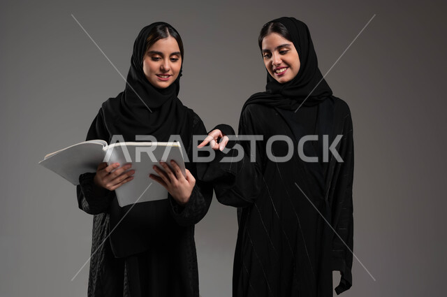 ثقافة التعلم الذاتي، مراجعة الدروس اليومية، الاستمتاع بممارسة هواية المطالعة، بورتريه مقرب لبنتين توءم عربتين خليجيتين سعوديتين محجبتين مبتسمتين تقضيان وقتهما بالقراءة والتثقف، شابة تشير بإصبع السبابة إلى الكتاب، خلفية رمادية