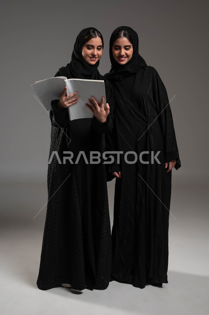 الاستمتاع بممارسة هواية المطالعة، قضاء الوقت بالقراءة والتثقف، بورتريه لبنتين توءم عربتين خليجيتين سعوديتين محجبتين تقرآان كتاب، دراسة ومراجعة الدروس اليومية، ثقافة التعلم الذاتي، صورة الجسم بالطول الكامل، خلفية رمادية