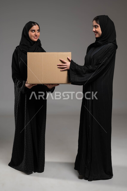 خدمات التوصيل المنزلي، استلام الطلبات الالكترونية، بورتريه لتوأم بنات عربيات خليجيات سعوديات يرتديان العباءة والحجاب تحملان صندوق مشتريات الاونلاين السريع، جودة شركات الشحن السعودية، صورة الجسم بالطول الكامل، خلفية رمادية