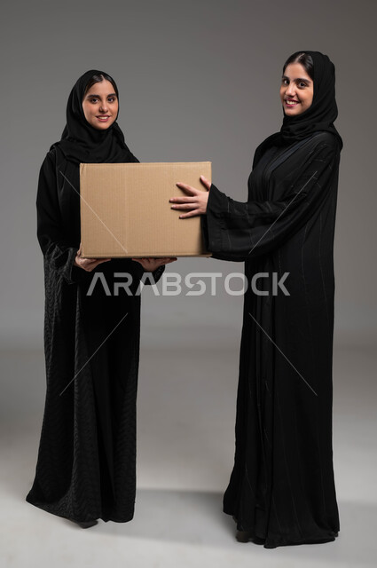Home delivery services, looking at the camera and receiving electronic orders, portrait of twin Saudi Gulf Arab girls wearing abayas and hijab carrying an express online shopping box, quality of Saudi shipping companies, full-length body photo, gray background