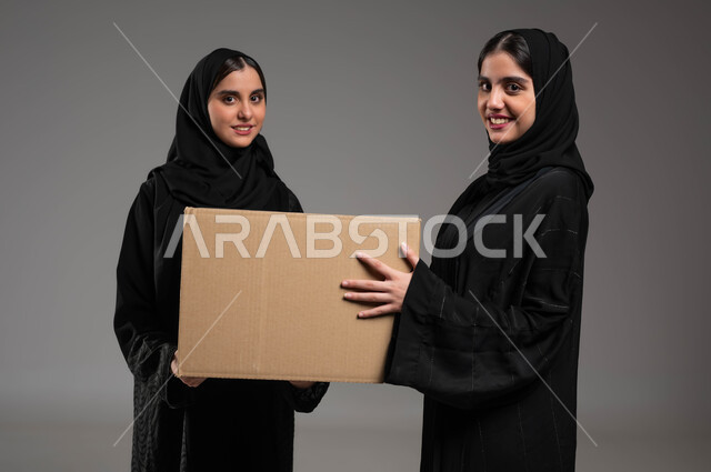 الوقوف والنظر إلى الكاميرا، جودة شركات الشحن السعودية، استلام الطلبات الالكترونية، بورتريه مقرب لتوأم بنات عربيات خليجيات سعوديات يرتديان العباءة والحجاب تحملان صندوق مشتريات الاونلاين السريع، خدمات التوصيل المنزلي، خلفية رمادية