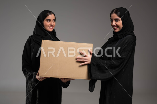 جودة شركات الشحن السعودية، استلام الطلبات الالكترونية، بورتريه مقرب لتوأم بنات عربيات خليجيات سعوديات يرتديان العباءة والحجاب تحملان صندوق مشتريات الاونلاين السريع، خدمات التوصيل المنزلي، خلفية رمادية