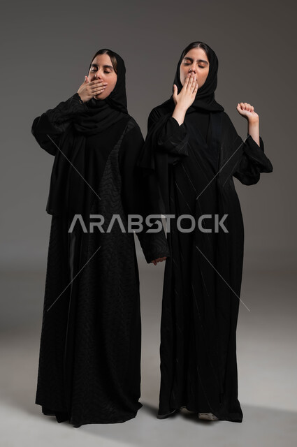النعاس والارهاق والحاجة إلى النوم, الإحساس بالكسل والخمول وطلب الراحة, بورتريه لتوأم بنات عربيات خليجيات سعوديات ترتديان العباءة السوداء والحجاب تقومان بالتثاؤب, الوقوف بتعب بإيماءات وضع اليد على الفم, السهر لوقت متأخر, صورة الجسم بالطول الكامل, خلفية رمادية