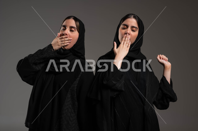 Standing tiredly with hand-to-mouth gestures, drowsiness, fatigue and need to sleep, feeling of laziness and lethargy and seeking rest, close-up portrait of twin Saudi Gulf Arab girls wearing the black abaya and hijab, yawning, staying up late, gray background