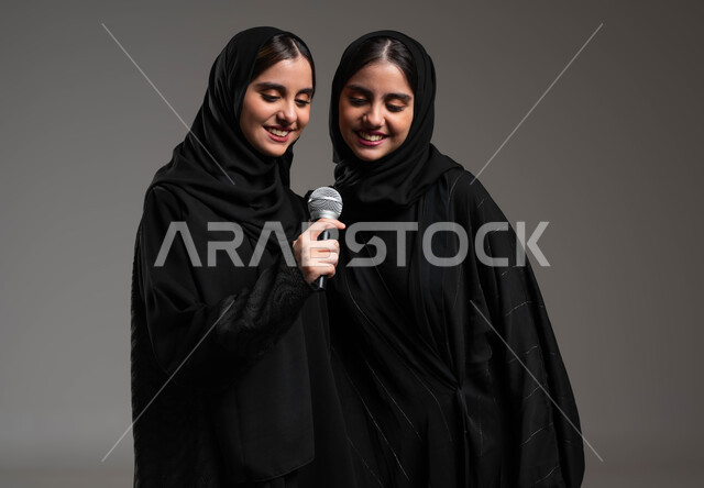 الدعم الفني للمواهب الغنائية في السعودية, الوقوف بخجل والاستمتاع بالغناء مع الأخوات, تسجيل المقاطع الصوتية باستخدام الميكرفون, بورتريه مقرب لتوأم بنات عربيات خليجيات سعوديات ترتديان العباءة والحجاب تقومان بتجربة الغناء المسرحي, خلفية رمادية