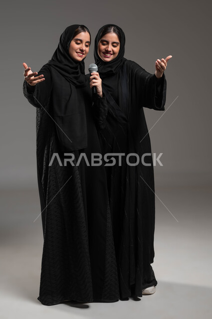 الاندماج والتفاعل بالغناء مع الأخوات, تسجيل المقاطع الصوتية باستخدام الميكرفون, الدعم الفني للمواهب الغنائية في السعودية, بورتريه لتوأم بنات عربيات خليجيات سعوديات ترتديان العباءة والحجاب تقومان بتجربة الغناء المسرحي, صورة الجسم بالكامل, خلفية رمادية