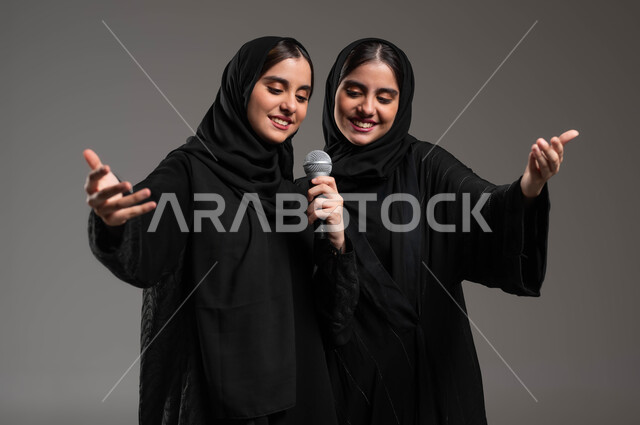 تقديم المواهب الصوتية بإيماءات الحماس والسرور, التفاعل والاندماج مع الموسيقى, مواهب فنية موسيقية, بورتريه مقرب لتوأم بنات عربيات خليجيات سعوديات يرتديان العباءة والحجاب تقومان بعرض غنائي مسرحي, الاستمتاع بممارسة هواية الغناء باستخدام الميكرفون, خلفية رمادية