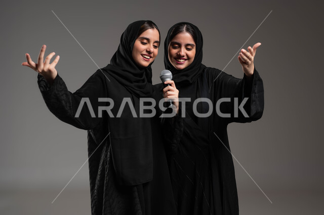 Practicing the hobby of singing using the microphone, interacting and merging with music, artistic musical talents, close-up portrait of twin Saudi Gulf Arab girls wearing the abaya and veil, performing a theatrical singing show, presenting vocal talents with gestures of enthusiasm and pleasure, gray background