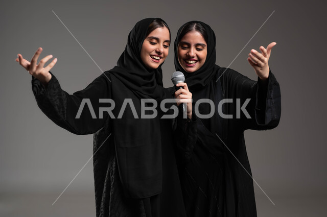 Interaction and merging with music, artistic musical talents, close-up portrait of twin Saudi Gulf Arab girls wearing abayas and hijab performing a theatrical singing performance, enjoying the hobby of singing using the microphone, presenting vocal talents with gestures of enthusiasm and pleasure, gray background