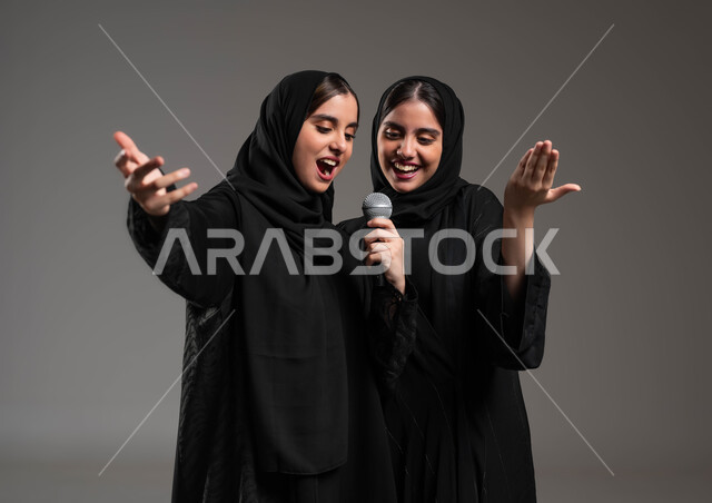 الاستمتاع بممارسة هواية الغناء باستخدام الميكرفون, تقديم المواهب الصوتية الفنية الموسيقية بإيماءات الحماس والسرور, بورتريه مقرب لتوأم بنات عربيات خليجيات سعوديات يرتديان العباءة والحجاب تقومان بعرض غنائي مسرحي, التفاعل والاندماج مع الموسيقى, خلفية رمادية