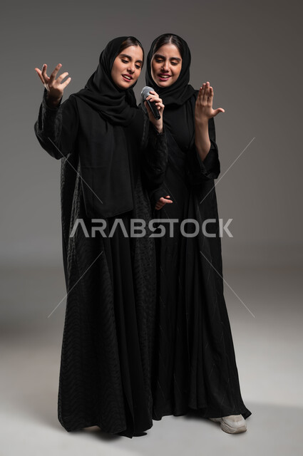 الدعم الفني للمواهب الغنائية في السعودية, التفاعل والاندماج بالغناء مع الأخوات, تسجيل المقاطع الصوتية باستخدام الميكرفون,  بورتريه لتوأم بنات عربيات خليجيات سعوديات ترتديان العباءة والحجاب تقومان بتجربة الغناء المسرحي, صورة الجسم بالكامل, خلفية رمادية