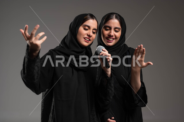 تقديم المواهب الصوتية الفنية الموسيقية بإيماءات الحماس والسرور, بورتريه مقرب لتوأم بنات عربيات خليجيات سعوديات يرتديان العباءة والحجاب تقومان بعرض غنائي مسرحي, التفاعل والاندماج مع الموسيقى, الاستمتاع بممارسة هواية الغناء باستخدام الميكرفون, خلفية رمادية