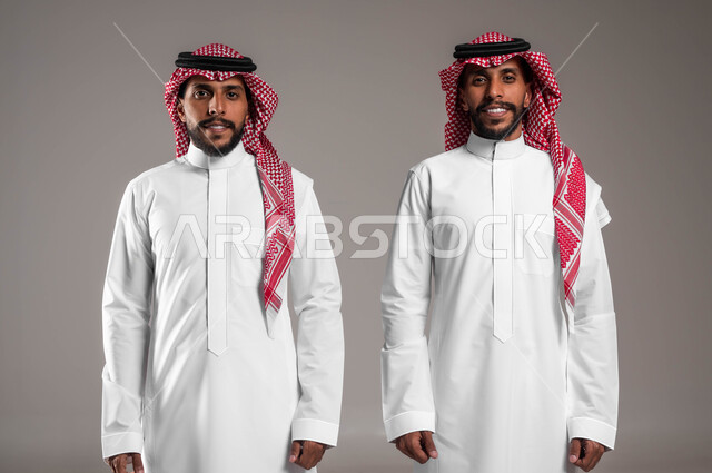 مفهوم الرجولة والأناقه، بورتريه لشابان توءم عربيان خليجيان سعوديان مبتسمان يرتديان الثوب التقليدي يقفان باستقامة وينظران إلى الكاميرا يإيماءات السرور، الاهتمام بالمظهر الخارجي، خلفية رمادية .