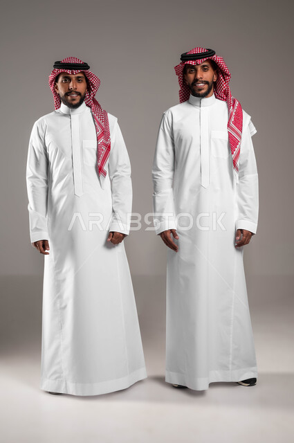 الاهتمام بالمظهر الخارجي، مفهوم الرجولة والأناقه، بورتريه لشابان توءم عربيان خليجيان سعوديان مبتسمان يرتديان الثوب التقليدي يقفان باستقامة وينظران إلى الكاميرا يإيماءات السرور، صورة الجسم بالطول الكامل، خلفية رمادية .