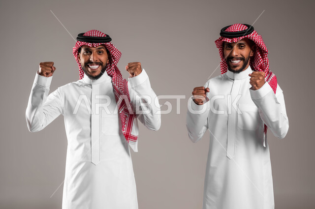 الشعور بالفرح والسعادة، مشاعر المحبة والود بين الاخوة، بورتريه لرجلان توءم عربيان خليجان سعوديان يرتديان الزي التقليدي وينظران الى الكاميرا بايماءات السرور، رفع اليدين عاليا بتعابير الفوز والنجاح وتحقيق الأهداف، خلفية رمادية