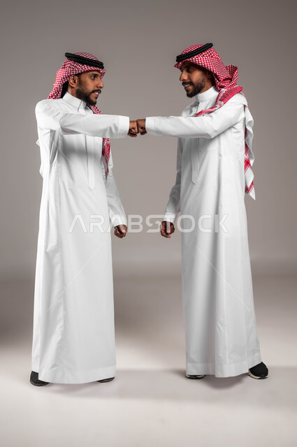 Distancing and following safety precautions to prevent the Corona virus, maintaining distances between people, a portrait of two young men, smiling Saudi Gulf Arab twins wearing traditional dress and shaking hands with a fist, full-length body portrait, gray background.