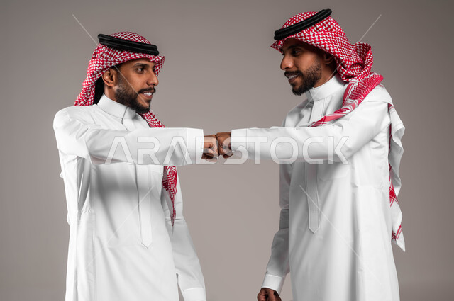 الحفاظ على المسافات بين الاشخاص، بورتريه لشابان توءم عربيان خليجيان سعوديان مبتسمان يرتديان الزي التقليدي و يقومان بالتصافح بقبضة اليد، التباعد واتباع احترازات السلامة  للوقاية من فايروس كورونا، خلفية رمادية