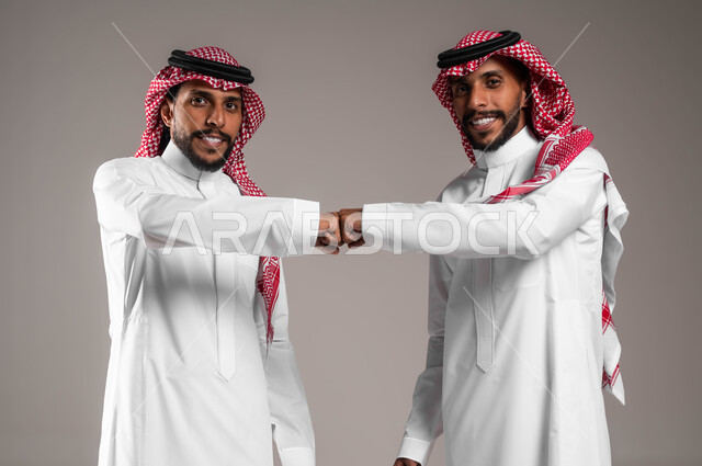 التباعد واتباع احترازات السلامة، الحفاظ على المسافات بين الاشخاص، بورتريه لرجلين توءم عربيين خليجيين سعوديين يرتديان الزي التقليدي و يقومان بالتصافح بقبضة اليد، الالتزام بإجراءات الوقاية من فايروس كورونا، النظر إلى الكاميرا يتعابير السرور، خلفية رمادية