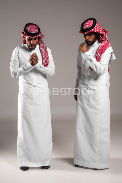 الإحساس بالبرد الشديد في الأجواء الشتوية، مفهوم التوءم الحقيقي المتطابق, بورتريه لشابين عربيين خليجيين سعوديين مبتسميين يرتديان الثوب والشماغ التقليدي يقفان بإيماءات تدل على الشعور بالصقيع والقشعريرة، صورة الجسم بالكامل، خلفية رمادية