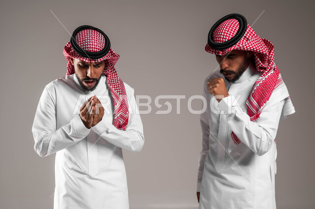 محاولة تدفئة الجسم، الإحساس بالبرد الشديد والقارس في فصل الشتاء, بورتريه لشابين توءم عربيين خليجيين سعوديين مبتسميين يرتديان الثوب والشماغ التقليدي يقفان بإيماءات تدل على الشعور بالصقيع والقشعريرة، خلفية رمادية