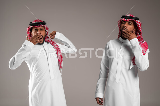 الشعور بالنعاس الشديد والرغبه في النوم، التعب والحاجة إلى الراحة بعد العمل, بورتريه لشابين توءم عربيين خليجيين سعوديين يرتديان الثوب والشماغ التقليدي يقفان بإيماءات التثاؤب، خلفية رمادية