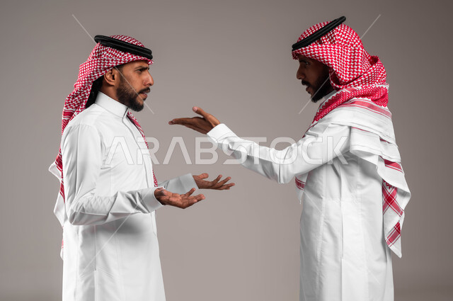 مفهوم النقاش الحاد والمواجهة بين الأخوة، بورتريه لشابين توءم عربيين خليجيين سعوديين يقفان مقابل بعضهما ويتجادلان بإيماءات الغضب وإلقاء اللوم، تعابير التعصيب والتوتر الانفعالي، خلفية رمادية