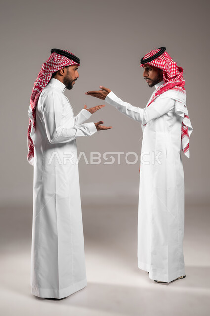 تعابير التعصيب والتوتر الانفعالي، مفهوم النقاش الحاد والمواجهة بين الأخوة، محاولة حل المشاكل العائلية، بورتريه لشابين توءم عربيين خليجيين سعوديين يقفان مقابل بعضهما ويتجادلان بإيماءات الغضب وإلقاء اللوم، صورة الجسم بالكامل، خلفية رمادية