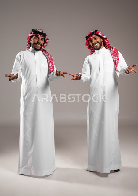 التردد وعدم الثقة بخصوص أمر معين، بورتريه لشابين توءم عربيين خليجيين سعوديين يرتديان الزي التقليدي يرفعان كفيهما بإيماءات عدم التأكد, الشعور بالحيرة وعدم المعرفة، الوقوف باستقامة والنظر إلى الكاميرا، صورة الجسم بالطول الكامل,خلفية رمادية