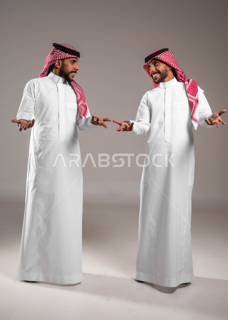الشعور بالحيرة وعدم المعرفة، التردد وعدم الثقة بخصوص أمر معين، بورتريه لشابين توءم عربيين خليجيين سعوديين يرتديان الزي التقليدي ينظران لبعضهما ويرفعان كفيهما بإيماءات عدم التأكد, صورة الجسم بالطول الكامل,خلفية رمادية