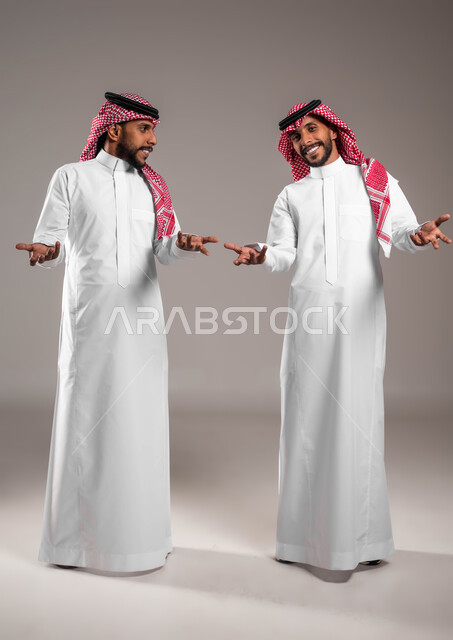 التردد وعدم الثقة بخصوص أمر معين، بورتريه لشابين توءم عربيين خليجيين سعوديين يرتديان الزي التقليدي يرفعان كفيهما بإيماءات عدم التأكد, الشعور بالحيرة وعدم المعرفة، الوقوف باستقامة والنظر إلى الكاميرا، صورة الجسم بالطول الكامل,خلفية رمادية
