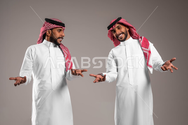 الوقوف باستقامة والنظر إلى الكاميرا، الشعور بالحيرة وعدم المعرفة، التردد وعدم الثقة بخصوص أمر معين، بورتريه لشابين توءم عربيين خليجيين سعوديين يرتديان الزي التقليدي يرفعان كفيهما بإيماءات عدم التأكد, خلفية رمادية
