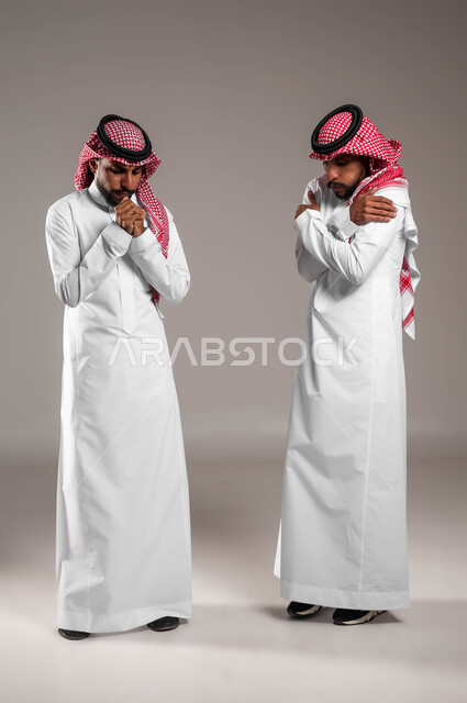 الإحساس بالبرد الشديد في الأجواء الشتوية، مفهوم التوءم الحقيقي المتطابق, بورتريه لشابين عربيين خليجيين سعوديين مبتسميين يرتديان الثوب والشماغ التقليدي يقفان بإيماءات تدل على الشعور بالصقيع والقشعريرة، صورة الجسم بالكامل، خلفية رمادية