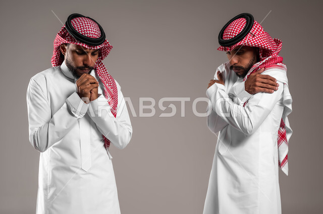 محاولة تدفئة الجسم، الإحساس بالبرد الشديد والقارس في فصل الشتاء, بورتريه لشابين توءم عربيين خليجيين سعوديين مبتسميين يرتديان الثوب والشماغ التقليدي يقفان بإيماءات تدل على الشعور بالصقيع والقشعريرة، خلفية رمادية