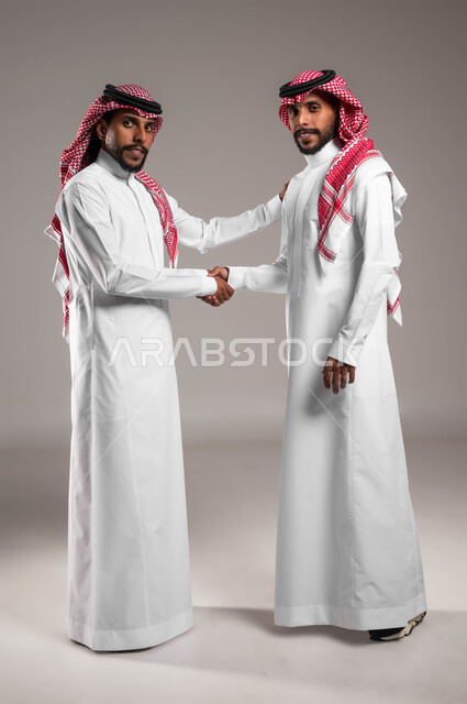 عقد صفقة واتفاقات، مفهوم التوءم الحقيقي المتطابق، النظر إلى الكاميرا بتعابير الترحيب، إلقاء التحية والسلام, بورتريه لشابين عربيين خليجيين سعوديين مبتسميين يرتديان الثوب والشماغ التقليدي يقومان بمصافحة بعضهما، صورة الجسم بالكامل، خلفية رمادية