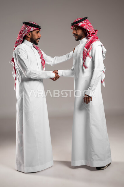 مفهوم التوءم الحقيقي المتطابق، عقد صفقة واتفاقات، الترحيب وإلقاء التحية والسلام, بورتريه لشابين عربيين خليجيين سعوديين مبتسميين يرتديان الثوب والشماغ التقليدي يقومان بمصافحة بعضهما، صورة الجسم بالكامل، خلفية رمادية