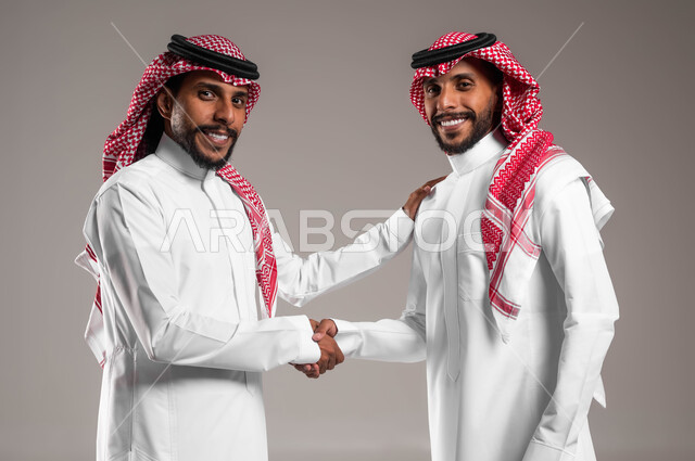 النظر إلى الكاميرا بإيماءات السرور والبشاشة والترحيب، عقد صفقة واتفاقات، إلقاء التحية والسلام, بورتريه مقرب لشابين توءم عربيين خليجيين سعوديين مبتسميين يرتديان الثوب والشماغ التقليدي يقومان بمصافحة بعضهما، خلفية رمادية
