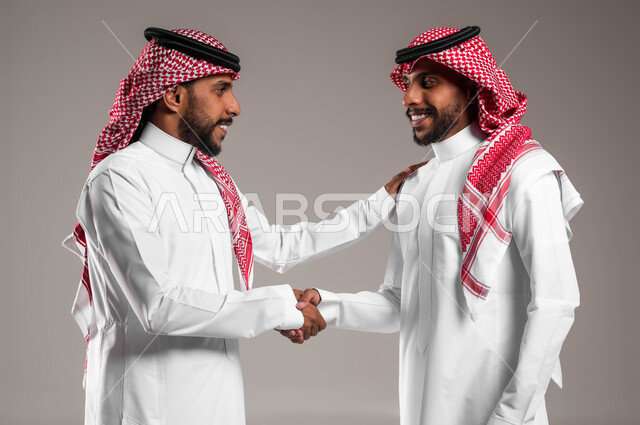 Making a deal and agreements, saying hello and greetings, expressions and gestures of happiness, cheerfulness and welcome, a close-up portrait of two smiling young Saudi Gulf Arab twins wearing the traditional thobe and shemagh, shaking hands with each other, gray background.