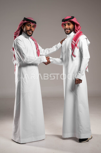 إلقاء التحية والسلام، عقد صفقة واتفاقات، تعابير وإيماءات السرور والبشاشة والترحيب،, بورتريه  لشابين توءم عربيين خليجيين سعوديين مبتسميين يرتديان الثوب والشماغ التقليدي يقومان بمصافحة بعضهما، صورة الجسم بالطول الكامل، خلفية رمادية