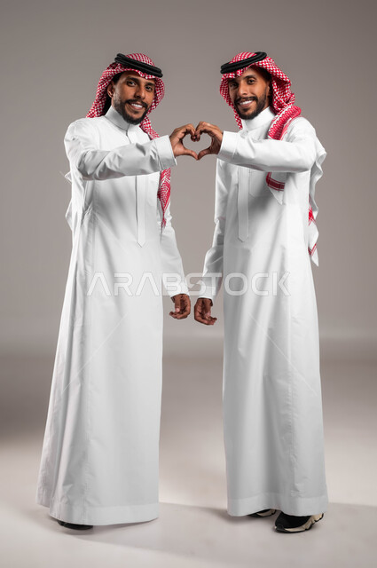 المحبة والمودة بين الأشقاء، بورتريه لرجلان توءم عربيين خليجيين سعوديين مبتسمين يرتديان الزي التقليدي يرفعان ايديهم ويشكلان قلب حب، النظر الى الكاميرا بتعابير السرور، تقوية وتعزيز الروابط العائلية، مفهوم الصداقة والاخوة، صورة الجسم بالكامل، خلفية رمادية