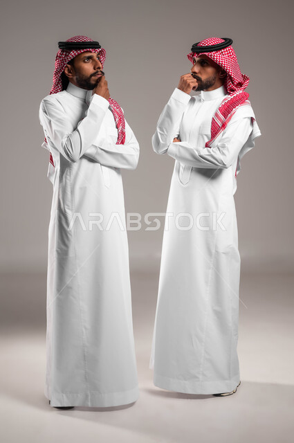 Planning for the future and being distracted, trying to find a solution to a problem, waiting for something with a bored expression, portrait of two Saudi Gulf Arab twin brothers wearing traditional clothing standing next to each other with thinking gestures, full-length body portrait, gray background.