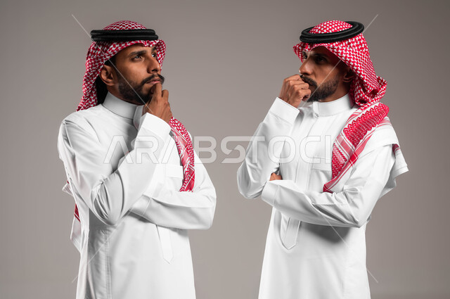 انتظار شيء ما بتعابير الملل، التخطيط للمستقبل وشرود الذهن، محاولة إيجاد حل لمشكلة ما، بورتريه لشقيقين توءم عربيين خليجيين سعوديين يرتديان الزي التقليدي يقفان بجانب بعضهما بإيماءات التفكير، خلفية رمادية .