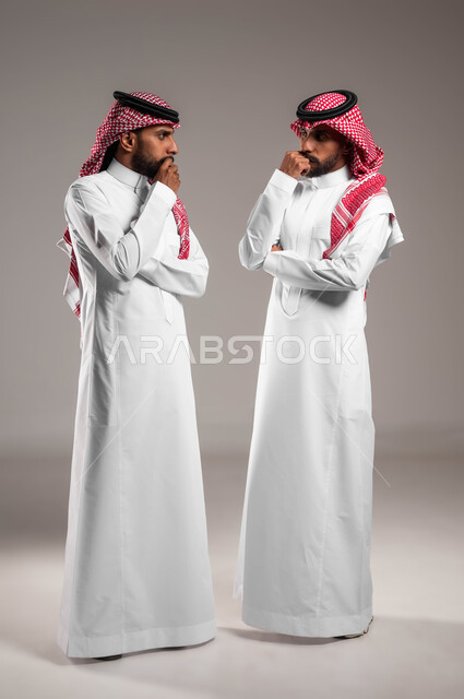 Absent-mindedness and contemplation, trying to find a solution to a problem, waiting for something with a bored expression, portrait of two Saudi Gulf Arab twin brothers wearing traditional dress and looking at each other with thinking gestures, full-length body portrait, gray background