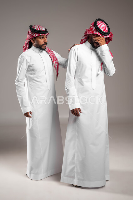 الشعور بالاكتئاب و الحزن، مفهوم التعاطف والمودة بين الأشقاء, الوقوف بجانب بعضهم البعض في الاوقات الصعبة, بورتريه لرجل عربي سعودي خليجي يرتدي الزي التقليدي يضع يده على كتف اخيه التوءم لتخفيف الالم والمواساة, صورة الجسم بالطول الكامل، خلفية رمادية