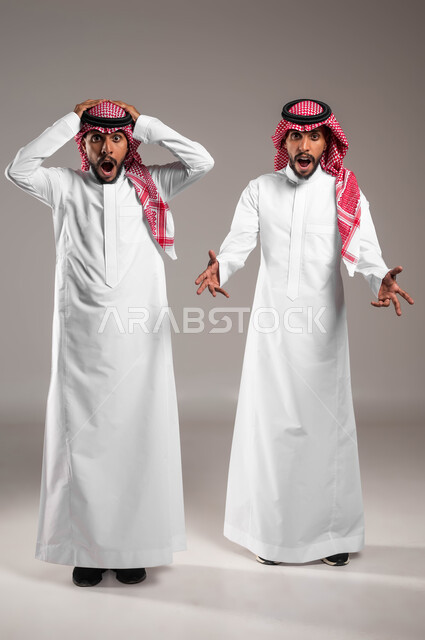 حركات وجه وفم تدل على الصدمة و الدهشة والذهول ,النظر إلى الكاميرا بإيماءات الذعر والخوف ,بورتريه لشابان توءم عربيان خليجيان سعوديان يرتديان الزي التقليدي و يرفعان أيديهم للأعلى بتعابير التفاجؤ عند رؤية شيء ما, صورة الجسم بالطول الكامل، خلفية رمادية .