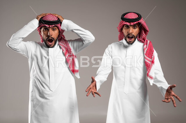 Looking at the camera with gestures of panic and fear, facial and mouth movements indicating shock, astonishment and astonishment, portrait of two young Saudi Gulf Arab twins wearing traditional clothing and raising their hands up with expressions of surprise when seeing something, gray background.