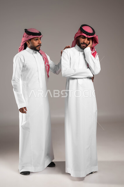 الشعور بالحزن و الاكتئاب، مفهوم التعاطف والمودة بين الأشقاء, الوقوف بجانب بعضهم البعض في الاوقات الصعبة, بورتريه لرجل عربي سعودي خليجي يرتدي الزي التقليدي يضع يده على كتف اخيه التوءم لتخفيف الالم والمواساة, صورة الجسم بالطول الكامل، خلفية رمادية