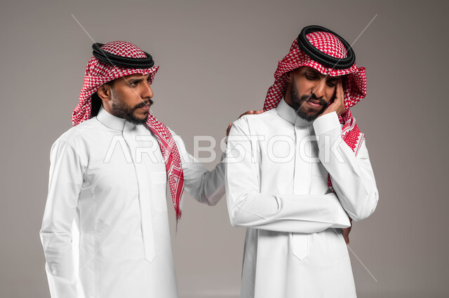 مفهوم التعاطف والمودة بين الأشقاء, الوقوف بجانب بعضهم البعض في الاوقات الصعبة, الشعور بالحزن و الاكتئاب، بورتريه لرجل عربي سعودي خليجي يرتدي الزي التقليدي يضع يده على كتف اخيه التوءم لتخفيف الالم والمواساة, خلفية رمادية