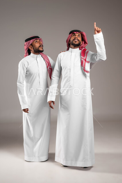 حركات الاستفسار والاشارة إلى شيء ما, رفع الاصبع عاليا بإيماءات تدل على وجود شيء ما, بورتريه مقرب لشابان توءم عربيان خليجيان سعوديان يرتديان الثوب التقليدي ينظران إلى الأعلى بتعابير الاستغراب و الذهول ،صورة الجسم بالطول الكامل، خلفية رمادية