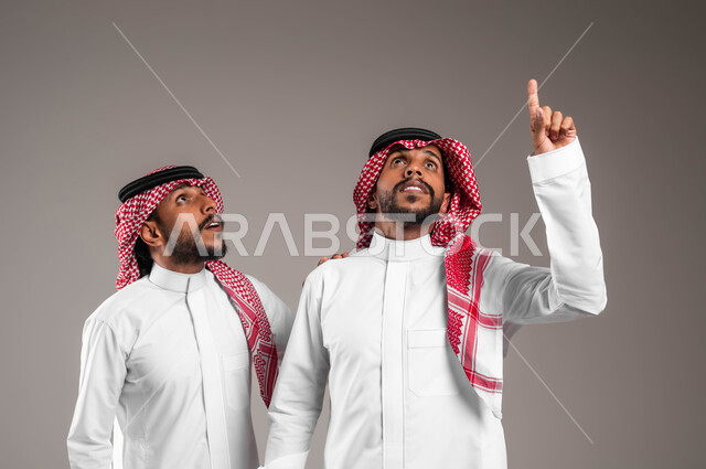 رفع الاصبع عاليا بإيماءات تدل على وجود شيء ما, حركات الاستفسار والاشارة إلى شيء ما, بورتريه مقرب لشابان توءم عربيان خليجيان سعوديان يرتديان الثوب التقليدي ينظران إلى الأعلى بتعابير الاستغراب و الذهول ،خلفية رمادية